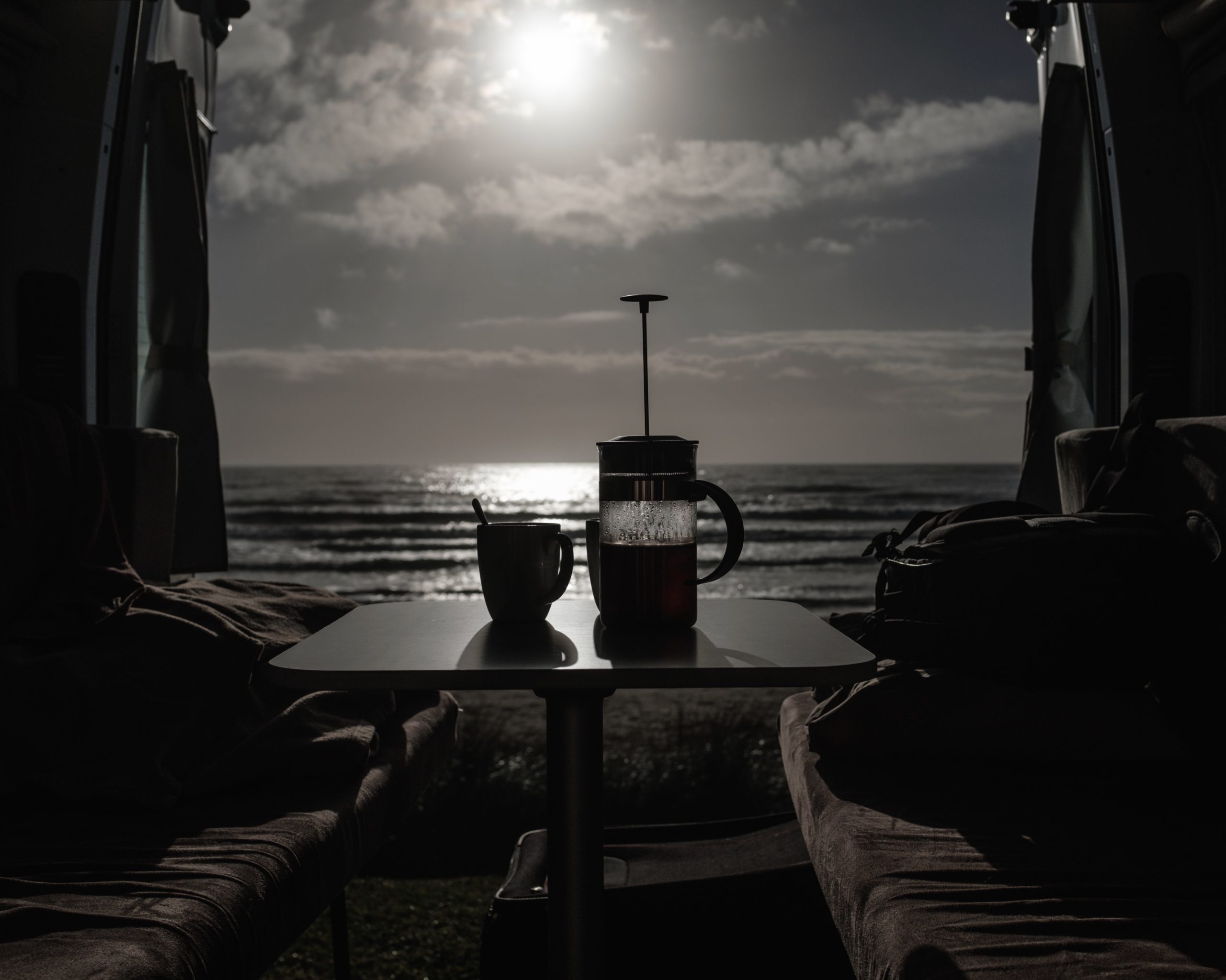 Depuis l'intérieur du campervan, durant la petit déjeuné. Une cafetière à piston et une tasse sont posées sur la table devant la porte arrière ouverte sur la plage au levé du soleil.